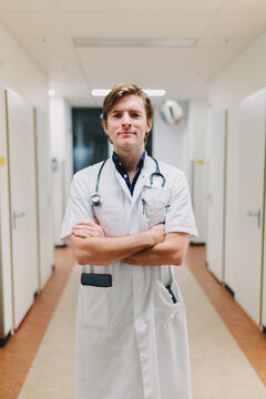 Young friendly doctor standing in the hall of a hospital with his arms crossed