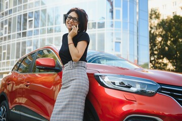 Beautiful businesswoman is talking on the mobile phone and smiling. She is standing near her car and looking forward with joy