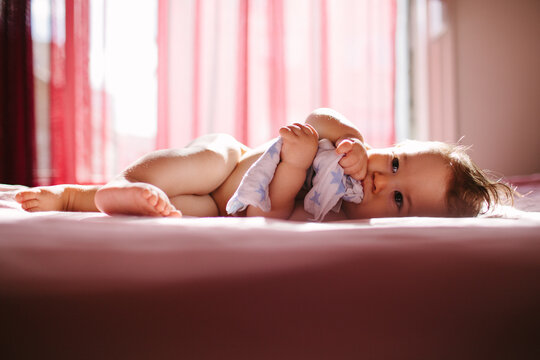 Baby holding a small blanket lies on a bed in beautiful light.