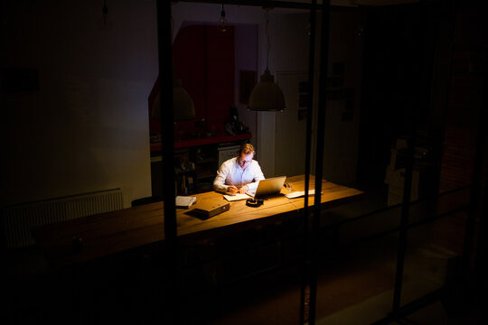 Young man working overtime in a dark office behind his laptop