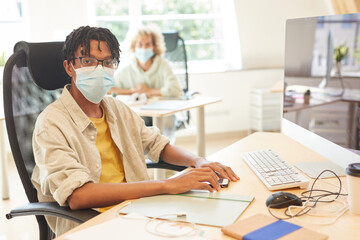 Portrait of African businessman in protective mask looking at camera while sitting at his workplace in front of computer monitor