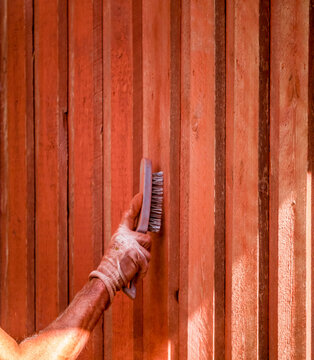 Hand is brushing old color of wood wall to prepair for new painting of red color on house outside.
