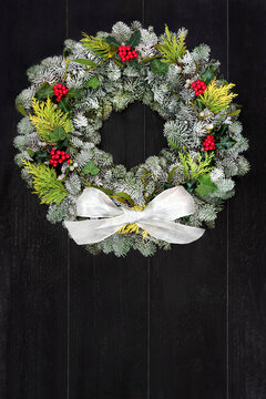 Traditional Christmas fir wreath with winter berry holly & greenery with white bow on rustic wood front door background. Festive composition for the solstice, Xmas & New Year holiday season. 