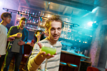 Caucasian young man looking at camera while posing with a cocktail in his hand and friends chatting, having drinks at the bar counter in the background