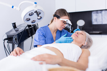 Professional cosmetician examining face skin of mature woman in medical esthetic office.