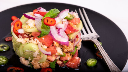 Italian tuscan vegetarian panzanella salad, mixed bread and vegetable salad on a black plate decorated with chilli peppers on a wooden light table, close-up