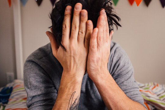 A man with tattoos sitting on a bed hiding his face in his hands out of frustration.