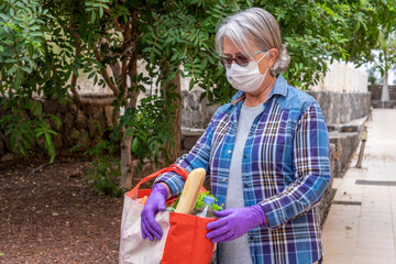 Senior woman people with gray hair comes back to home after shopping of groceries wearing surgical mask and protective gloves due to coronavirus pandemic