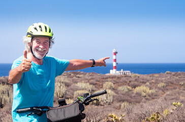 Cheerful bearded senior man with yellow helmet enjoying excursion with bicycle close to a lighthouse - arid landscape and horizon over water in a sunny day in Tenerife, canary islands