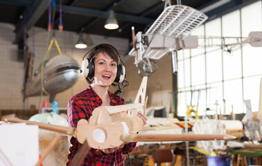 Portrait of happy female hobbyist in aviator headphones with unfinished monoplane model in aircraft workshop..