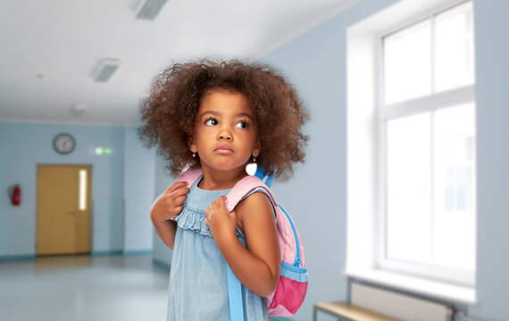 Childhood And Education Concept - Sad Little African American Girl With Backpack Over School Corridor On Background