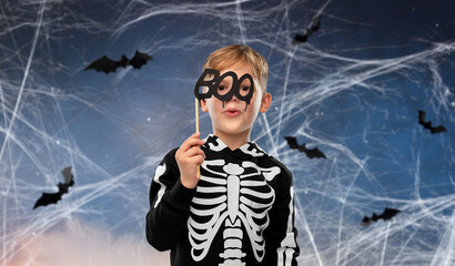 halloween, holiday and childhood concept - smiling boy in black costume of skeleton with party accessory making faces over night sky with bats and cobweb on background