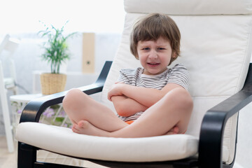 Offended little child boy sitting on couch entertaining in living room. Sad small kid enjoying free leisure time at home.