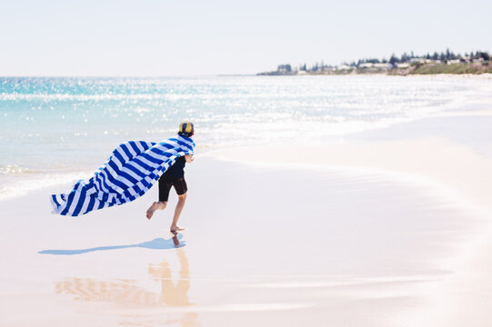 Boy wearing his towel as a cape while running on a beach beside a sparkly ocean on a sunny day
