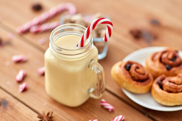 christmas and seasonal drinks concept - eggnog in glass mug with candy cane decoration, cinnamon buns and aromatic spices on wooden background