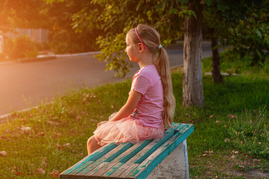 Cheerful little girl with long blonde hair in pink tulle skirt sitting alone on a bench on empty street on sunset. Happy dreamy cute child on nature background. Kids dress fashion. Greeting card.