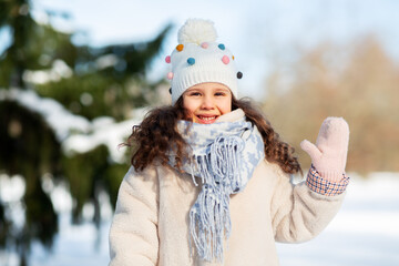 childhood, leisure and season concept - portrait of happy little girl in winter clothes waving hand at park