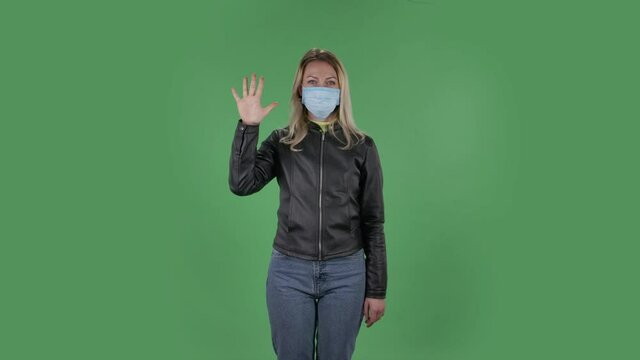 Portrait of beautiful young woman in medical protective face mask is looking at camera and shows fist, then counting one, two, three, four, five fingers. Blonde with loose hair in a black jacket and
