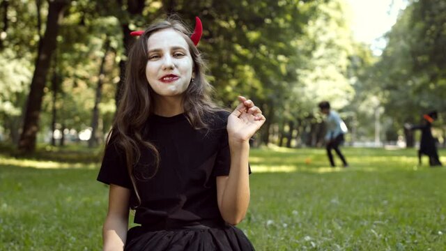 Cheerful Caucasian girl wearing devil costume is sitting on grass in park, taking sweets out of pumpkin buckets and eating them while other kids playing on background