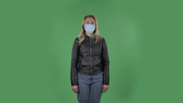Portrait of beautiful young upset woman in medical protective face mask is looking at camera gets nervous and then calms down and meditates. Blonde with loose hair in a black jacket and jeans on a