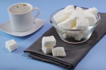 Pile of white homemade marshmallows in a transparent bowl on a blue background. Cup of hot coffee and homemade marshmallows. Food concept.