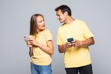 Portrait of lovely couple with smart phones in hands, cheerful charming woman checking her email, man follows her secret correspondence with jealous expression over grey background