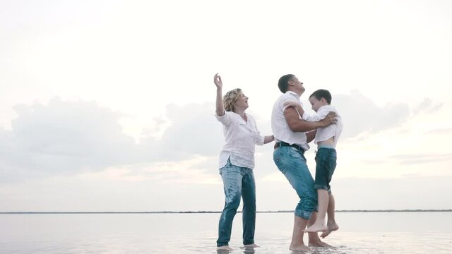 Loving father throws up little cheerful son, laughing mother embraces happy family standing in lake. Carefree couple with kid boy together plays funny flying games on vacation, summer leisure outdoors