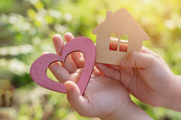 children's hands hold a wooden house and a heart, top view. The idea is peace and quiet in a house where love reigns. Soft focus, horizontal photo, close-up.