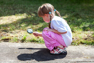 Little curious girl searching for something with a magnifying glass in the backyard. Young child squatting, looking for something on the ground with a loupe. Children curious about the world concept