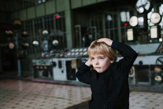 Boy dances with his hands on his head in front of the control panel of a disused power station.