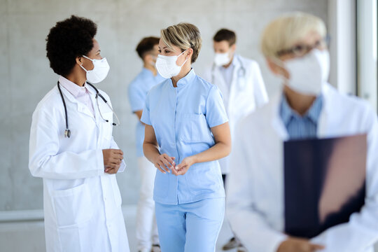 African American Doctor And Nurse Wearing Face Masks While Talking In A Hallway At Clinic.