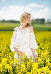 Naklejka na ściany i meble Happy girl posing in rape seed flowers field posing in white dress at sunny day outdoor