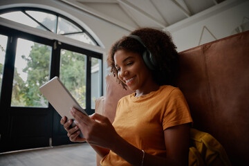 Cheerful young woman sitting on floor with backrest on couch using digital tablet and headphone