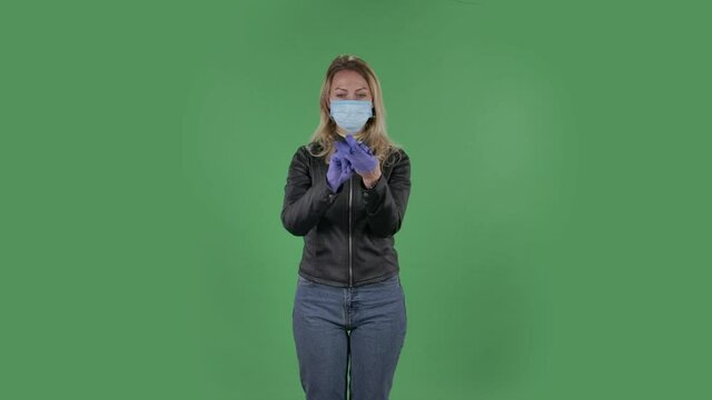 Portrait of beautiful young woman in medical mask is looking at camera and puts on rubber gloves. Blonde with loose hair in a black jacket and jeans on a green screen in the studio. Health Protection