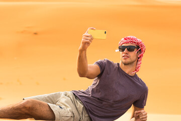 Young male tourist taking a selfie picture with his mobile phone while lying on a sand dune of the Arabian desert. Travel and tourism.