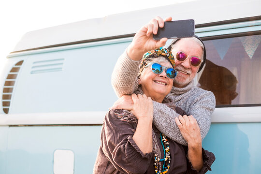 Cheerful old senior people couple hug and take a selfie picture together enjoying the outdoor leisure activity - travel concept with blue vintage van in background - happy retired elderly people