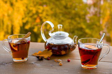 Autumn tea party outdoors. Glass kettle with hot herbal tea and two glass cups on a wooden table against the background of yellow trees. 