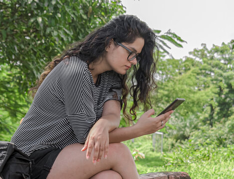 Beautiful and attractive young girl with open curly hair indulged deeply in her phone, updating her social media status, as she enjoys some quality leisure time in nature with lush green background.