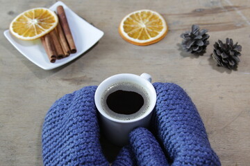 Autumn or winter cozy composition. Cup of coffee, cinnamon sticks, sugar, orange and pine cones on a wooden background. Hands in blue mittens holding a cup of coffee..