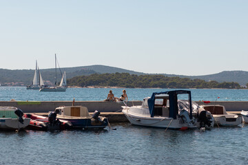 Fototapeta premium Biograd Croatia August 2020 Old small fishing boats docked in the stone harbour of Biograd, women sitting on the wall and sunbathing. Simple warm summer day on the sea