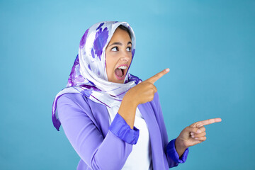 Young beautiful arab woman wearing islamic hijab over isolated blue background surprised, looking and pointing side