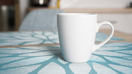 Clean white mug with handle stands on blue table behind blurred kitchen background