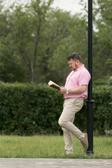 A man in a pink shirt with a book in his hands stands near a lighting pole.