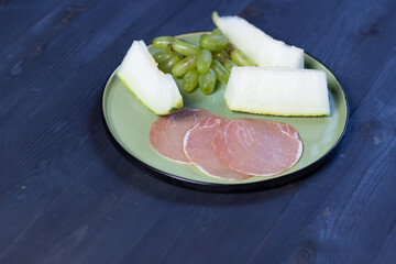 sliced prosciutto and melon, grape  on green  plate on black wooden background