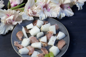 Gray Plate with melon and prosciutto on black wooden background and white gladiolus flowers...