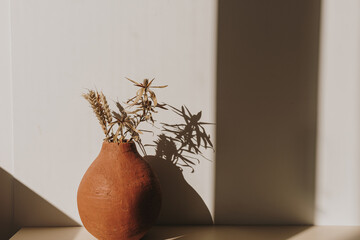 Red handmade clay flower pot with dry wheat / rye bouquet in sunlight shadows on white background....
