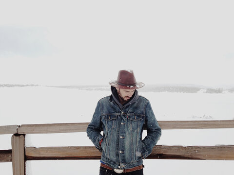 Handsome man leaning against fence with snow and hat