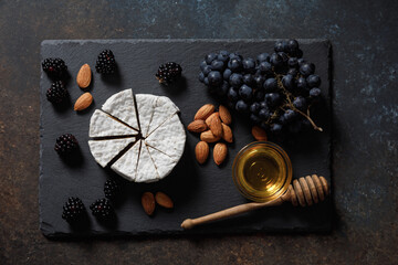 Camembert or brie french soft cheese with grapes, honey, nuts and berries on black stone serving board.
