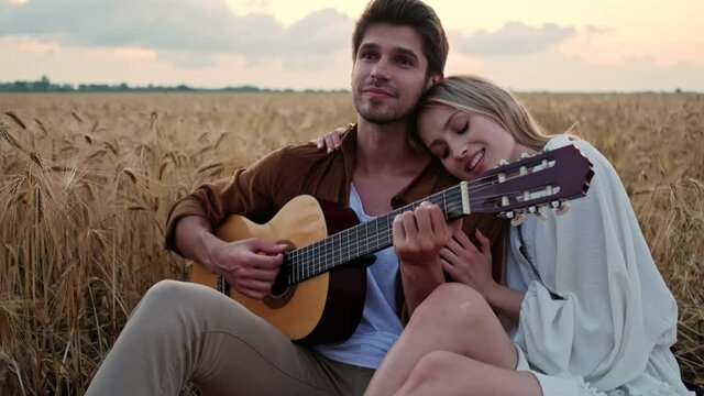 A young beautiful couple in love man and woman are sitting together while playing the guitar in the field