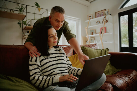 Wife And Husband Working From Home Looking At A Laptop On The Couch - Family Working From Home Looking On A Laptop While Doing Online Shopping 